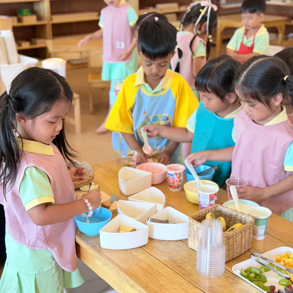 Children in aprons spooning yoghurt and fruit into bowls at a wooden table